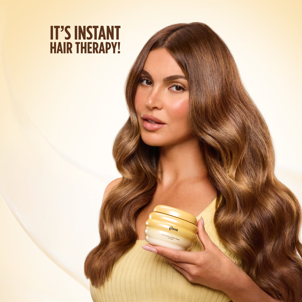 Model with long wavy brunette hair in a yellow top holding the hair mask against a glowing background.