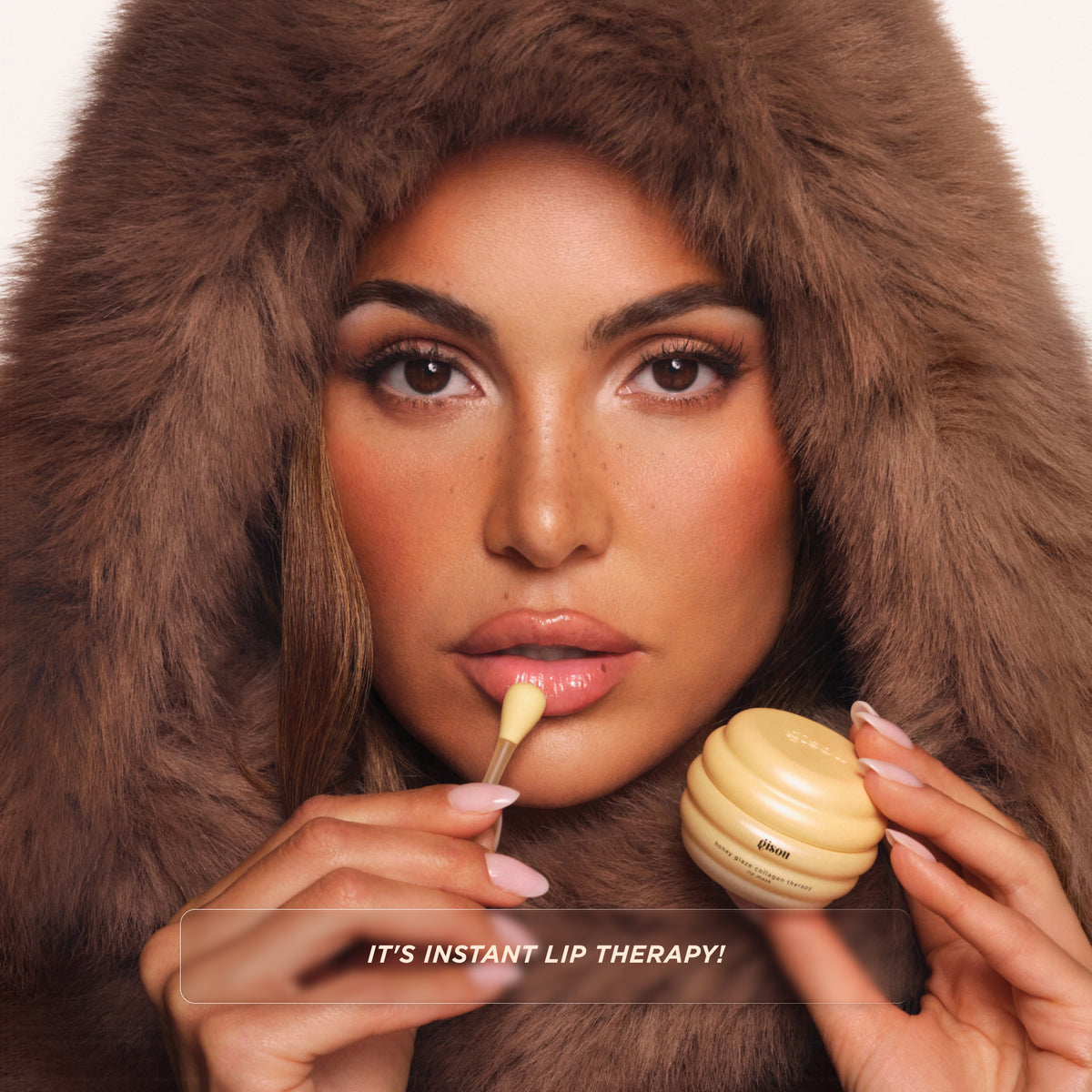 Model in a fur hood applying the Gisou Honey Glaze Collagen Therapy Lip Mask with an applicator.