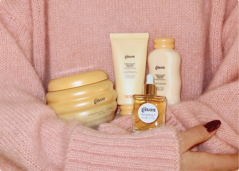 Gisou Honey Infused Hair Mask, haircare bottles and Honey Infused Hair Oil held against a pink knit sweater