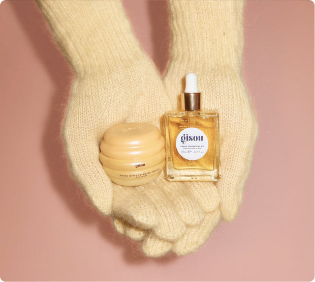 Hands in cream knit mittens holding Gisou Honey Infused Hair Oil and a hair mask on pink