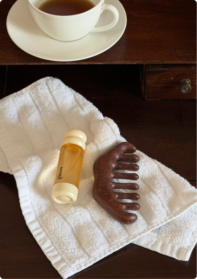 Gisou hair oil and wooden scalp massager on a white towel beside a cup of tea