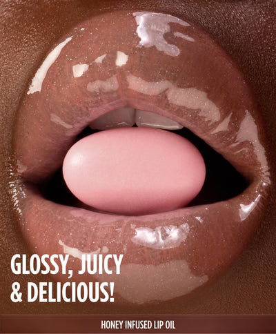 Close-up of glossy lips holding a pink candy between them, labeled 'Glossy, Juicy & Delicious!' for the Gisou Honey Infused Lip Oil.