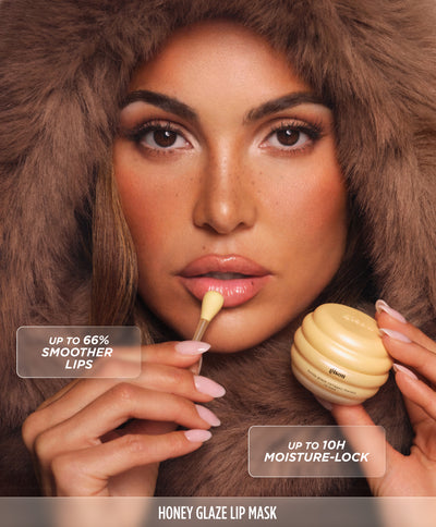 Model in a fur hood applying the Gisou Honey Glaze Collagen Therapy Lip Mask with an applicator.