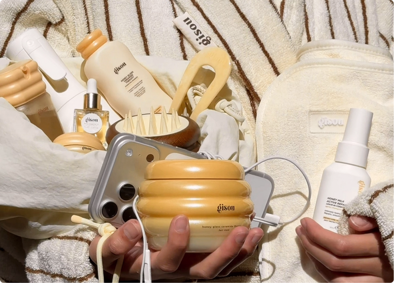 Hands holding Gisou's Honey Gloss Ceramide Therapy heated hair mask alongside the Honey Milk Active Repair Leave-In Conditioner, Honey Infused Hair Oil, and a yellow hair pick, styled with a striped Gisou robe and Honey Glow Hair towel.