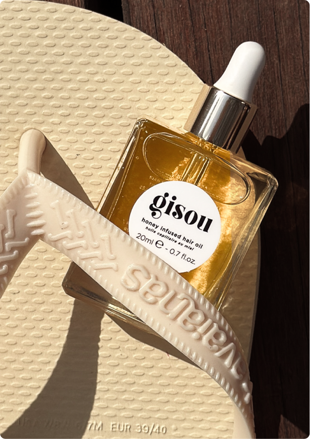 A bottle of Gisou Honey Infused Hair Oil resting on a cream flip-flop in warm sunlight.