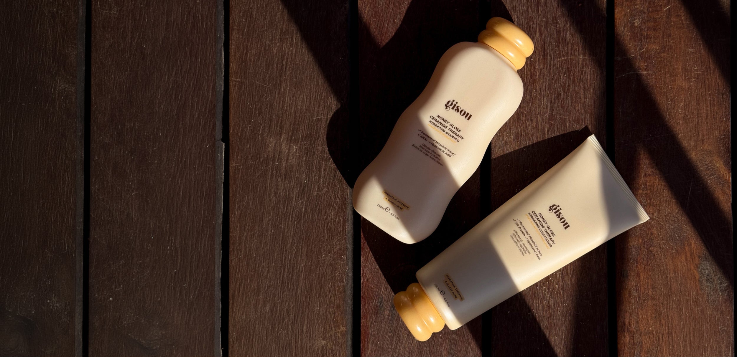 Gisou Honey Gloss Ceramide Therapy Hydrating Shampoo and Conditioner bottles lying on a sunlit wooden deck.