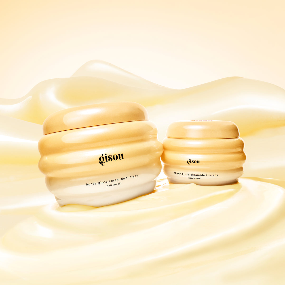 Two jars of Gisou Honey Gloss Ceramide Therapy Hair Mask in honey-inspired packaging placed on a glossy, golden, honey-like surface.