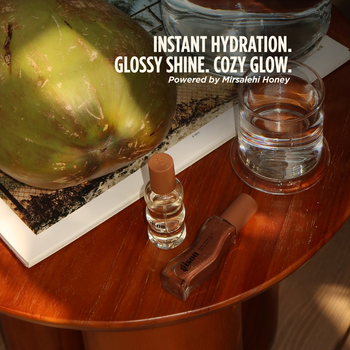 Warm lifestyle flat lay featuring a Coco-Cacao themed Gisou pairing on a polished wooden table. A mini Honey Infused Hair Perfume and shimmering cocoa-brown Honey Infused Lip Oil are styled beside a glass of water, a large green coconut and an open book in golden sunlight. Overlay text reads: “Instant hydration. Glossy shine. Cozy glow. Powered by Mirsalehi Honey.”