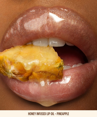 Close-up of glossy lips wearing Gisou Honey Infused Lip Oil in the Pineapple shade with golden shimmer, holding a fresh pineapple slice between the lips. The image highlights the high-shine finish and juicy tropical-inspired look of the lip oil.