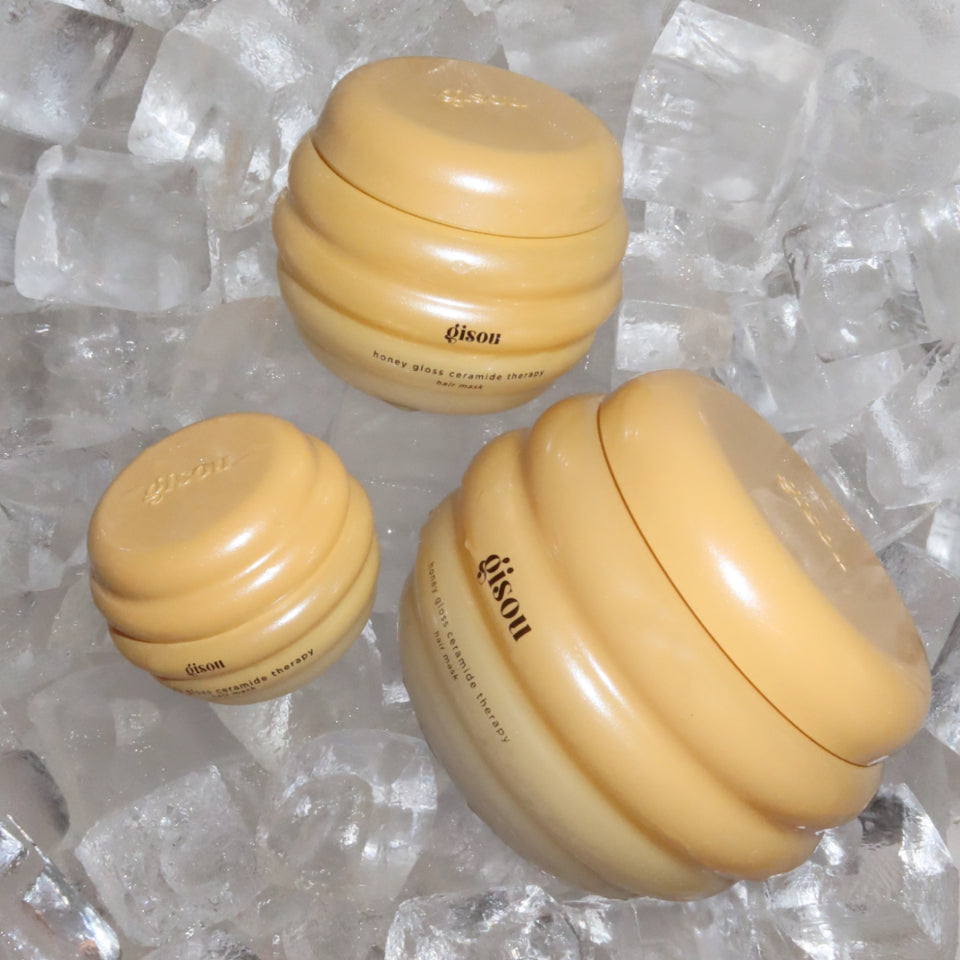 Three jars of Gisou Honey Gloss Ceramide Therapy Hair Mask in honey-inspired packaging placed on ice.