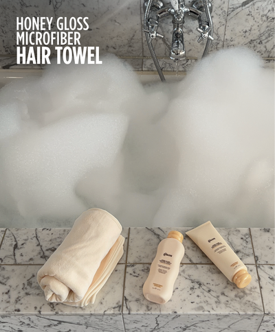 Gisou Honey Gloss Microfiber Hair Towel next to Shampoo and Conditioner laying near the bath full of foam.