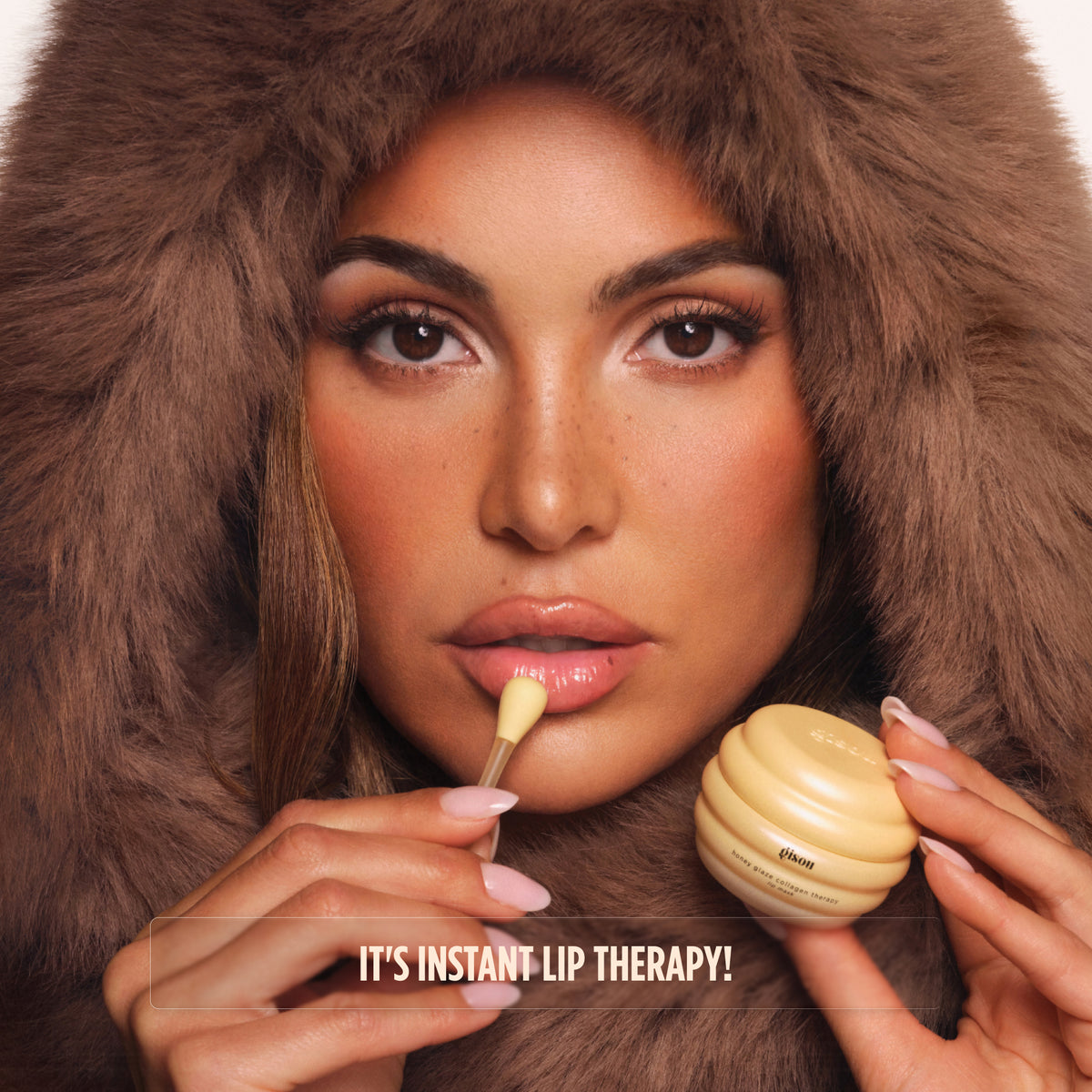 Model in a faux fur hood applying Gisou lip mask: It's instant lip therapy!